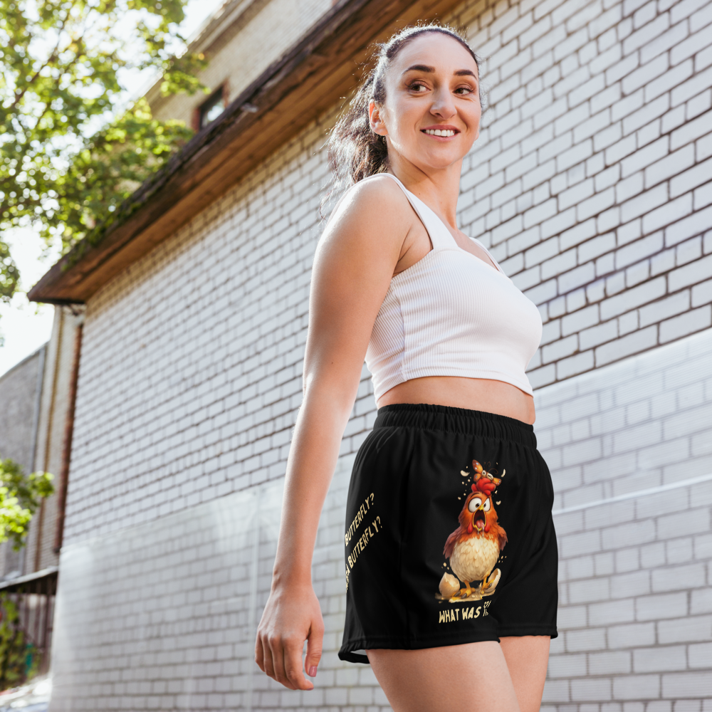 Woman wearing a white crop top and black shorts with a rooster graphic, standing against a brick wall.
